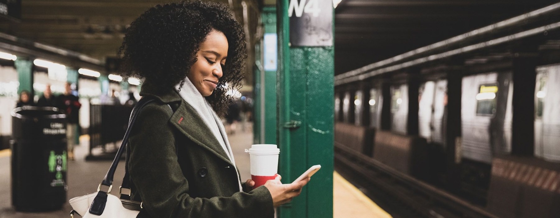 woman with a cup of coffee texts on subway station platform